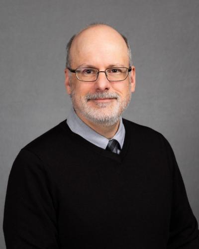A man wearing a black sweater, collared shirt, and tie, with glasses and a goatee, against a gray background.