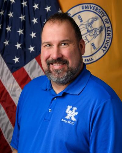 A man smiles and poses in front of a University of Kentucky flag and an American flag.