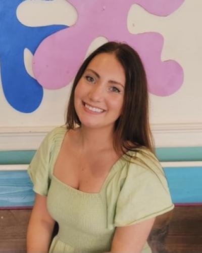 A woman with long brown hair smiles while sitting in front of a colorful wall.